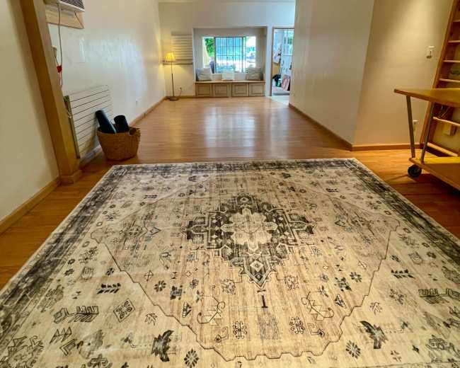 The image shows a spacious, well-lit room featuring a large decorative rug, with a staircase on the right and a bright area visible in the background.