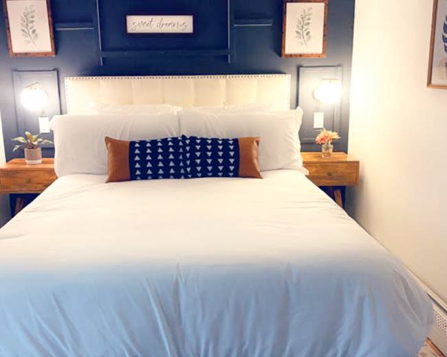 The image shows a neatly arranged bedroom with a white bedspread, decorated with a dark blue accent pillow, flanked by wooden nightstands and framed botanical prints on the wall.