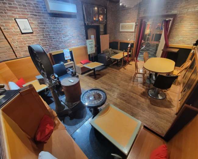 The image shows a cozy room with exposed brick walls, wooden furniture arranged around a central table, and a mix of seating, including chairs and benches.