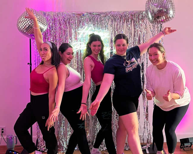 Five women pose in front of a shiny silver backdrop with disco balls, dressed in athletic wear and striking playful poses.