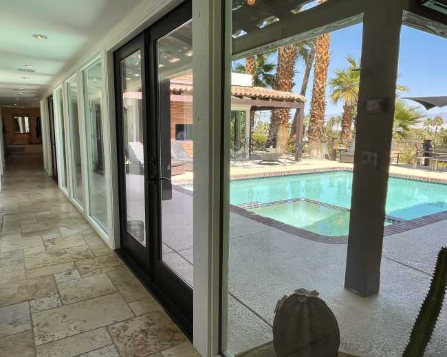 The image shows a hallway with large glass doors leading to a patio with a swimming pool and palm trees in the background.
