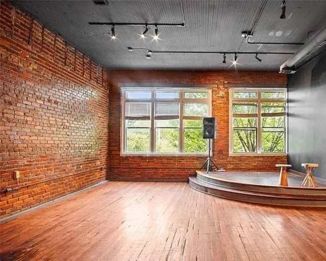 The image shows an empty room with exposed brick walls, large windows, a wooden stage, and two stools.