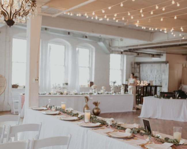 The scene shows a well-decorated event space with long tables set for a gathering, featuring white tablecloths, centerpieces, candles, and string lights overhead.