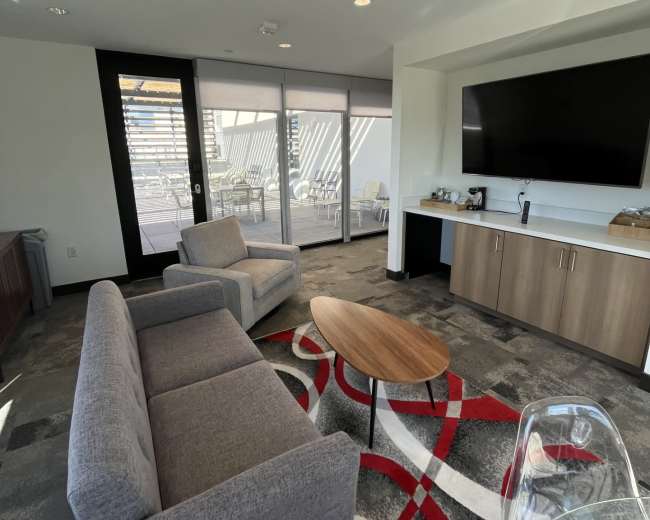 The image shows a modern lounge area featuring two gray sofas, a wooden coffee table, and a television mounted on the wall, with large windows providing natural light.