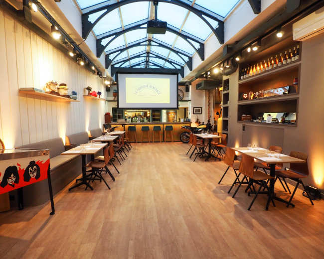 The image shows a modern restaurant interior with wooden floors, tables arranged for dining, a large screen on the wall, and shelves displaying various decorations.