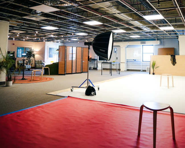 The image shows a spacious studio with a red mat, a stool in the center, lighting equipment, and partitions creating distinct areas.