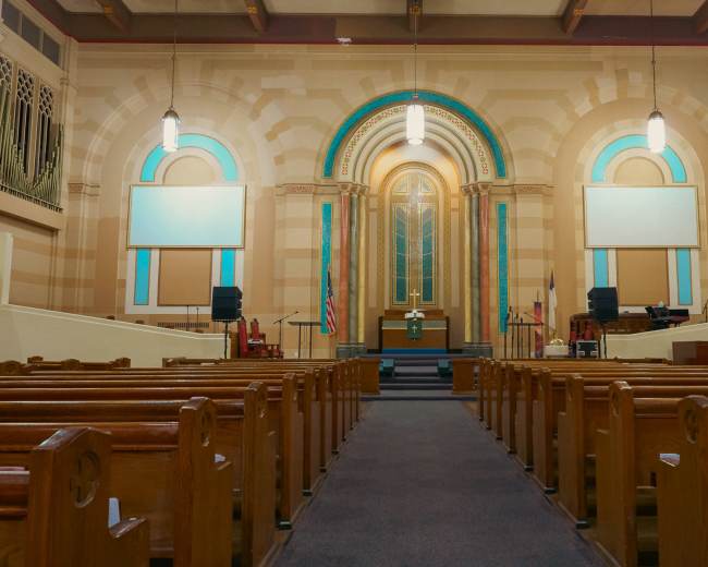 The image shows the interior of a church with wooden pews facing a raised altar area, illuminated by hanging lights and large windows.