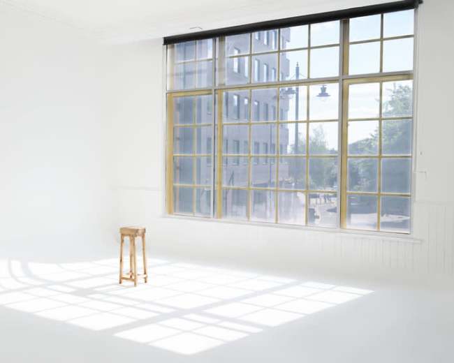 A wooden stool sits in a bright, empty room beside a large window with a grid pattern.