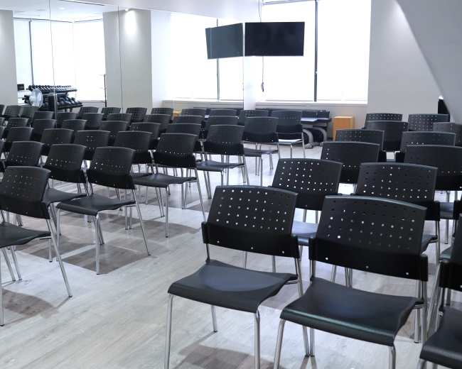 The image shows a spacious room arranged with rows of black, stackable chairs facing a blank television screen and large windows.