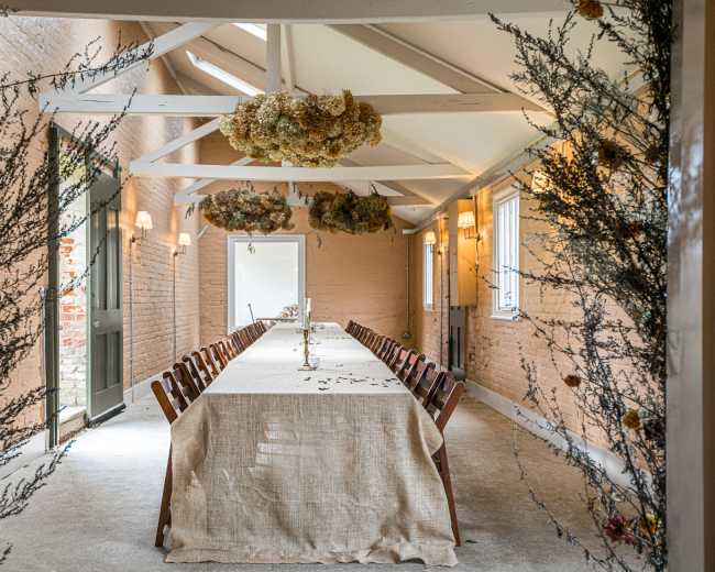 A long dining table is set in a rustic room, adorned with dried floral decorations and natural lighting from large windows.
