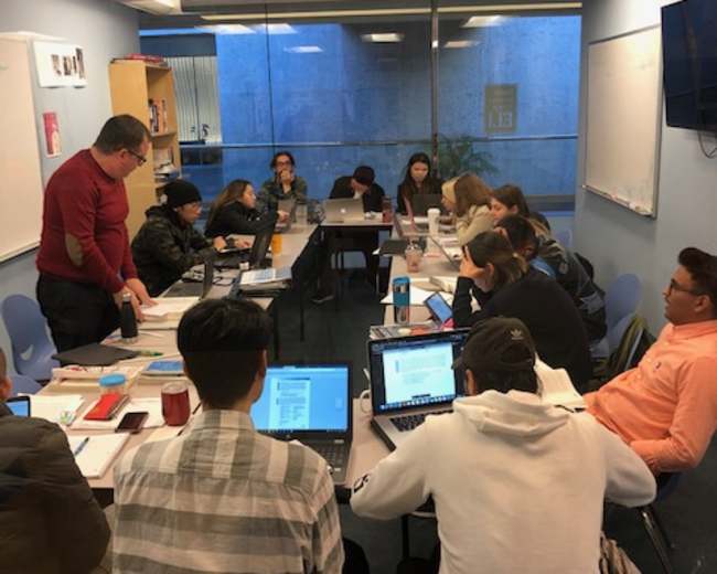 A group of students and a teacher are seated around a table in a classroom, engaged with laptops and study materials.