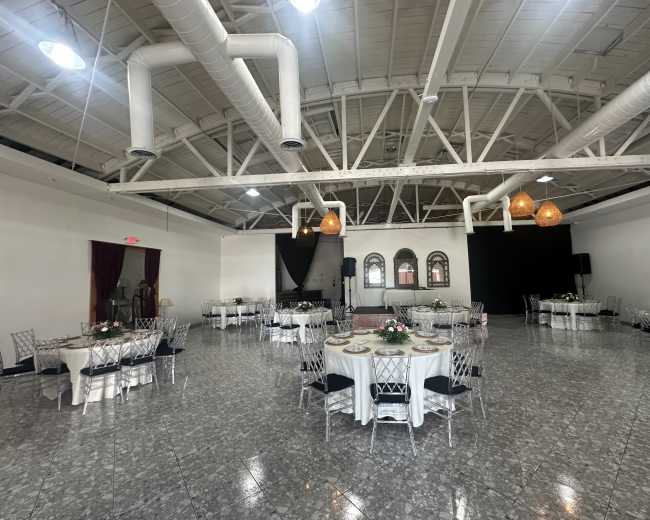 The spacious interior features several round tables set with white tablecloths, arranged under a high ceiling with exposed beams and large windows.