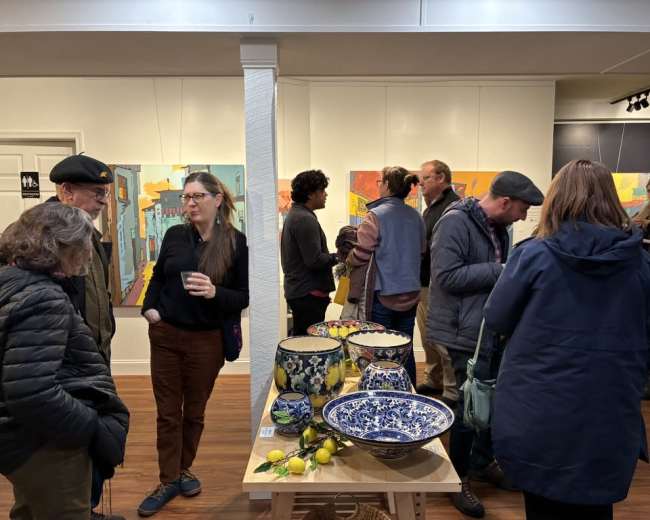 The scene depicts a gallery opening with people engaging in conversation around art displays and a table adorned with decorative bowls and fruit.