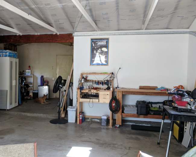 The image shows a garage with a workbench, a fan, and various tools on one side, while a doorway leads to another room in the background.