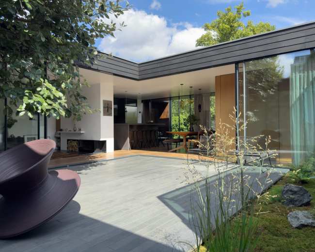 The image shows a modern home with a central courtyard featuring a sculptural seating piece, surrounded by glass walls and greenery.