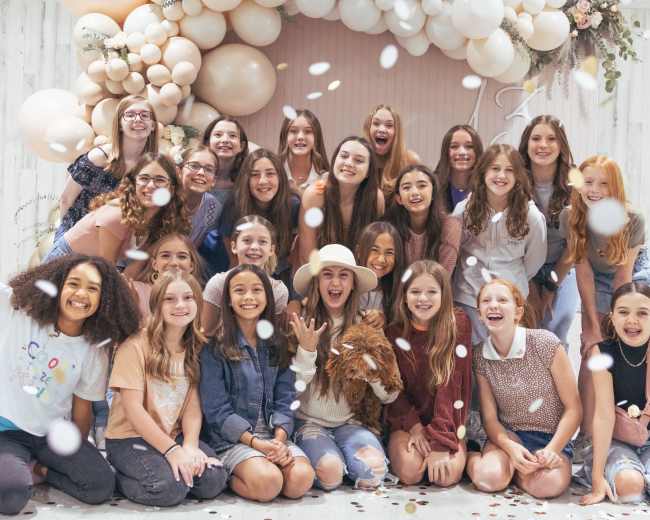 A group of young girls poses together in front of a decorative balloon backdrop, with confetti falling around them.