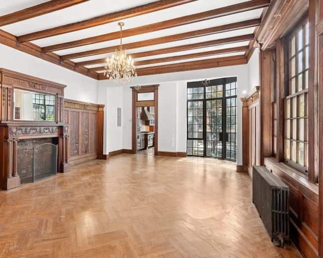 The image shows a spacious interior room featuring wooden beams on the ceiling, a decorative fireplace, and large windows that illuminate the space.