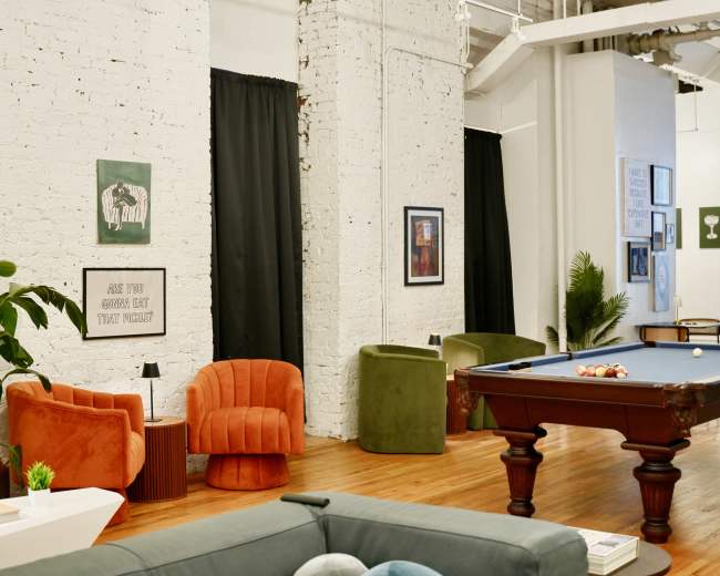 The image shows a spacious interior featuring a billiard table, colorful seating areas, and decorative wall art against white brick walls.