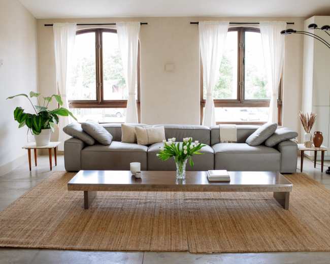 A modern living room features a gray sofa with pillows, a wooden coffee table, and large windows with sheer curtains, complemented by potted plants and a floor lamp.