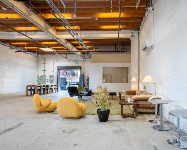 The image shows an open industrial-style interior space with a mix of seating areas, including a yellow lounge chair and sofas, and tables set up against the walls.