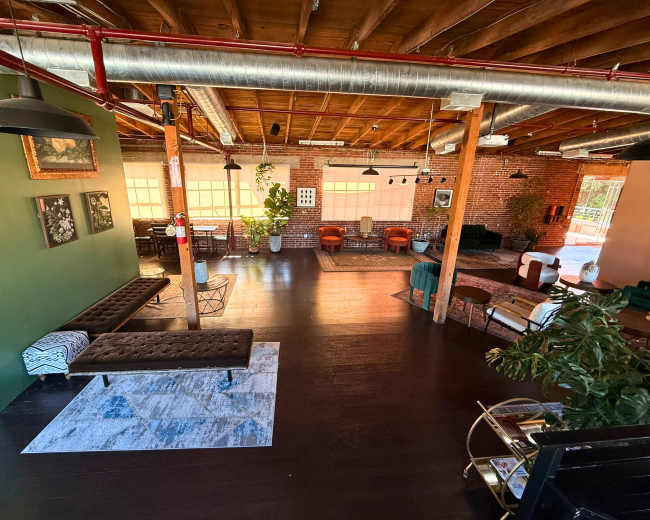 The image shows a spacious, modern interior with wooden beams and a mix of seating arrangements, including couches and chairs, surrounded by large windows and green walls.