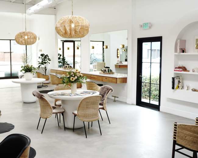 The image shows a modern, minimalist interior space featuring a round dining table surrounded by chairs, pendant lighting, and large windows with plants visible outside.