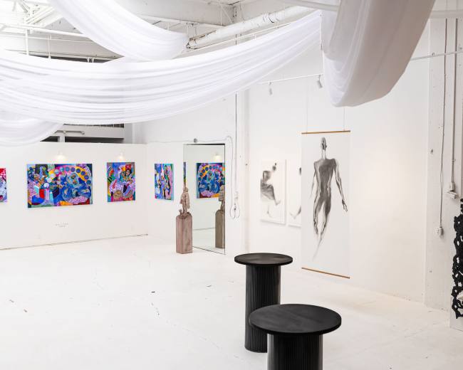 The image shows an art gallery featuring colorful paintings on the walls, a sculpture in the center, and two black round tables.