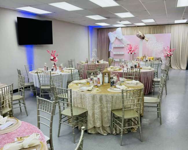 The image shows a banquet hall set up for an event, featuring round tables covered with tablecloths and centerpieces, and decorated with pink accents.