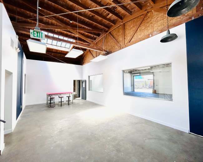 The image shows a spacious, well-lit room with a high ceiling, featuring exposed beams, concrete flooring, and a table with stools.