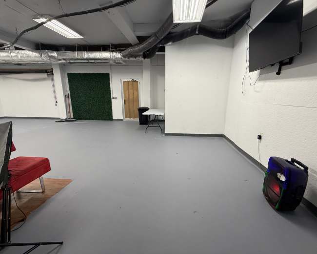 The image shows a spacious room with gray flooring, a television mounted on the wall, a table in the corner, and a green wall decoration.