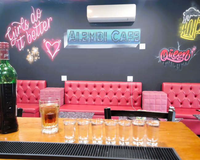 The image shows a bar with a wooden countertop featuring several shot glasses lined up in front of a glass of a dark beverage and a bottle, with a colorful mural on the wall behind it.