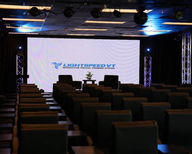 The image shows a seminar room set up with rows of chairs facing a large presentation screen, featuring the "LIGHTSPEED VT" logo and two chairs on a stage.