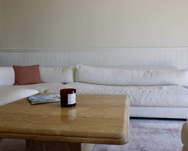 A light-colored sofa sits across from a marble-top coffee table adorned with a candle and magazines in a bright room.