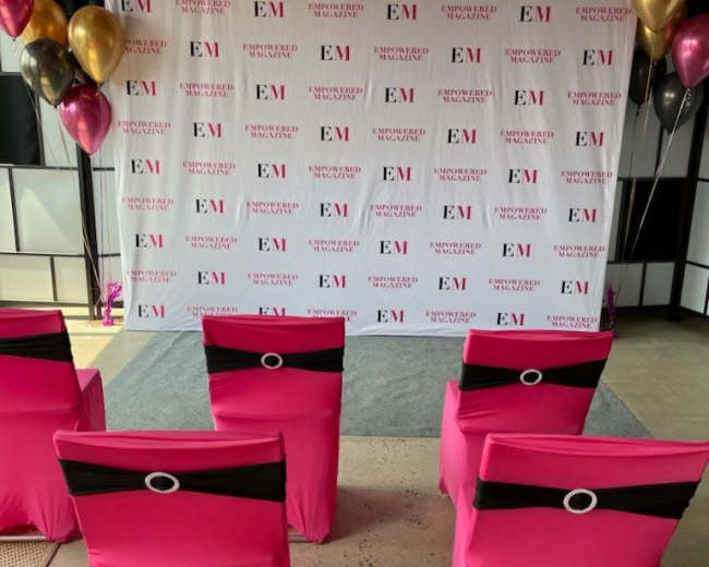 The image shows a decorated event space with pink chair covers and a backdrop featuring the letters "EM" and branded designs.