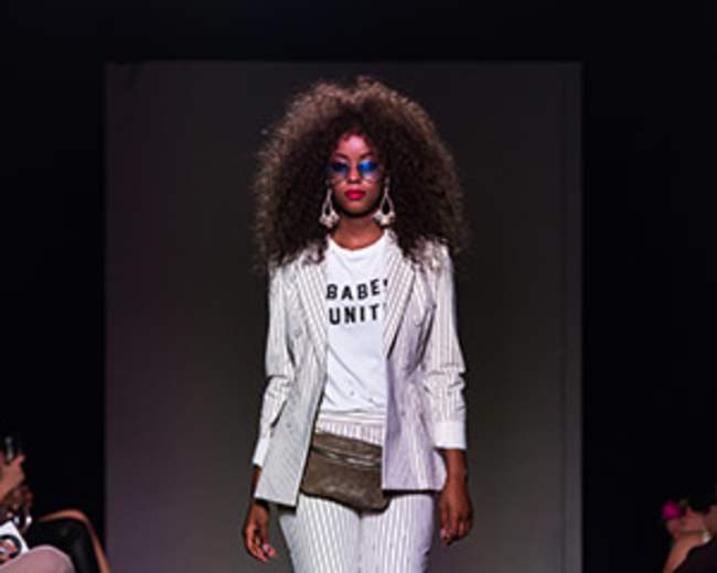 A model walks down a runway wearing a striped suit over a graphic t-shirt, with an audience seated on either side.