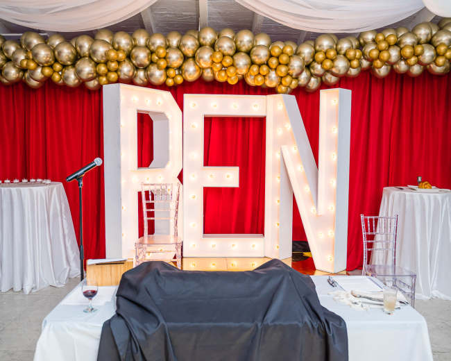 The scene features large illuminated letters spelling "REN" against a backdrop of red curtains and a decorative arrangement of gold and silver balloons.