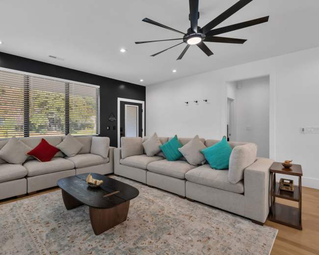 A modern living room features a light-colored sectional sofa, a dark wooden coffee table, and large windows with blinds, surrounded by a neutral color palette.