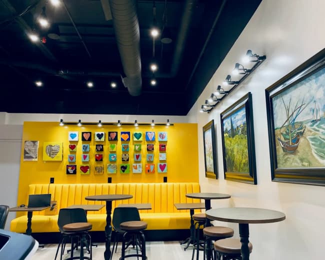The image shows a modern cafe interior featuring yellow walls adorned with colorful artwork and a seating area with a yellow bench and circular tables.