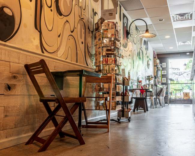 The image shows a narrow corridor of a café lined with artwork on the walls and displaying various items for sale on a stand.