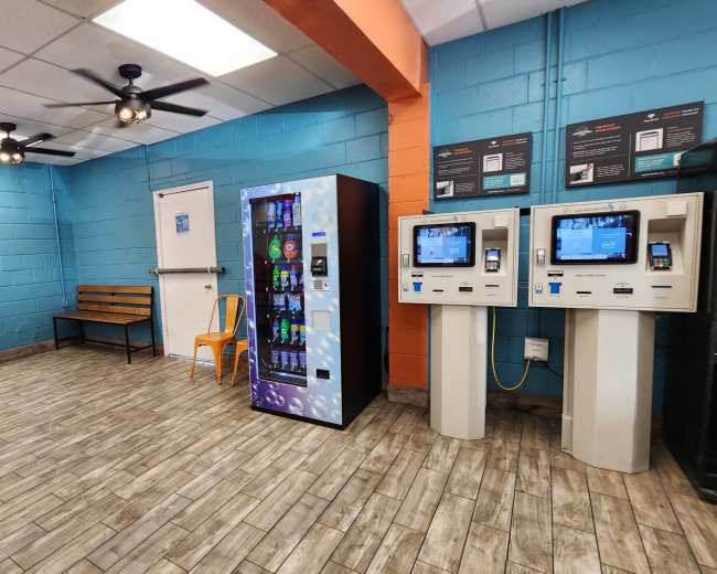 The image shows an empty laundromat with a vending machine, two self-service kiosks, and laundry machines along the walls.