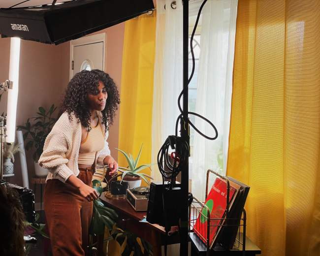 A woman with curly hair stands by a wooden table, adjusting a turntable surrounded by plants and vinyl records in a brightly lit room with yellow curtains.