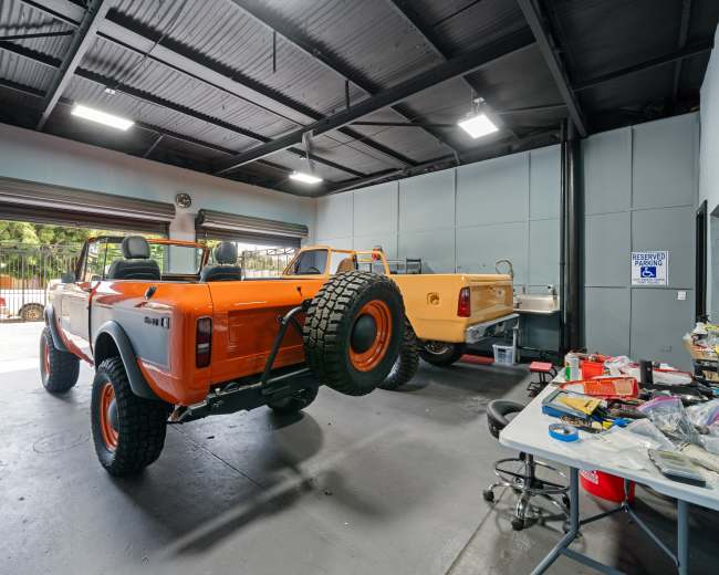 The image shows an orange off-road vehicle in a garage alongside a yellow vehicle, with various tools and equipment scattered on a table.