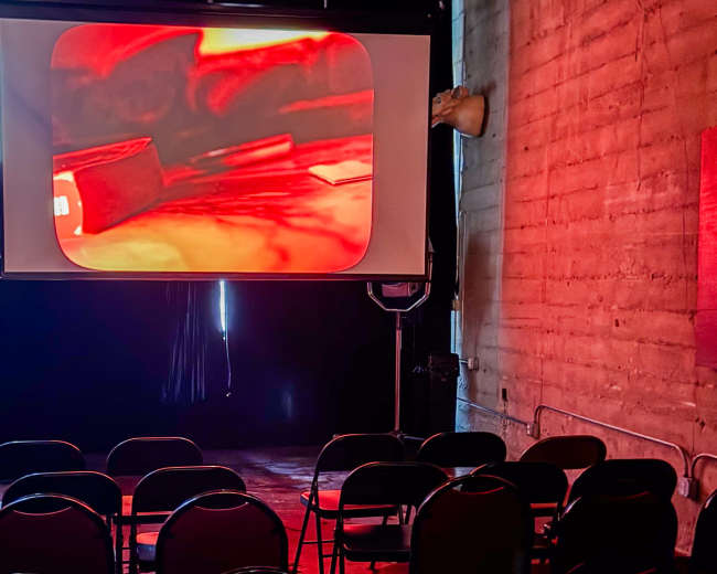 A projection screen displays a red-tinted image in a dimly lit room with empty black chairs arranged in front.
