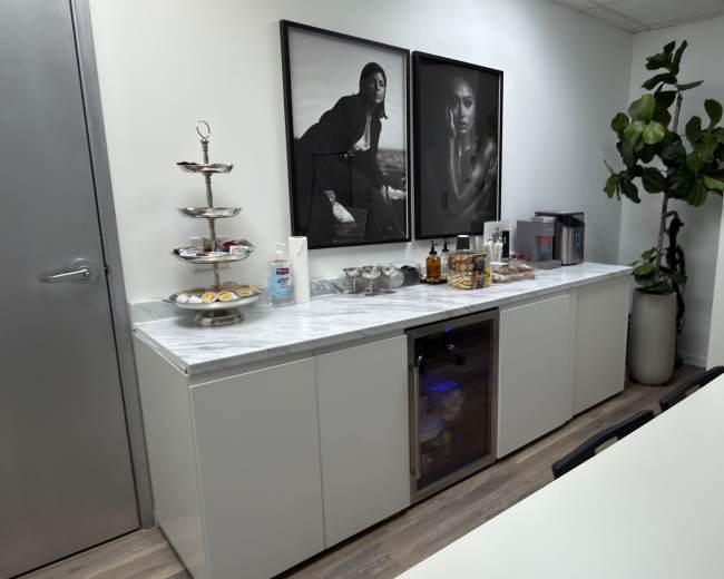 The image shows a modern office kitchenette with a marble countertop, a tiered serving stand with pastries, a mini bar with alcoholic beverages, two framed black-and-white photographs on the wall, and a large potted plant.