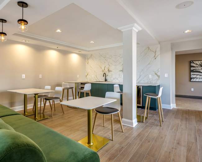 A modern, well-lit lounge area featuring green sofas, sleek tables with gold legs, and a kitchen counter with marble accents.