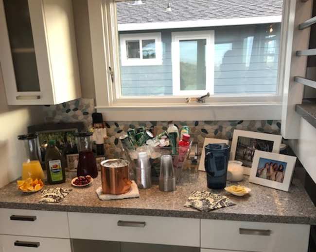 A kitchen countertop with various beverages, snacks, and a framed photo, set against a window overlooking a house exterior.