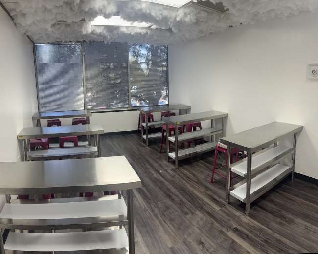 The image shows a classroom with multiple metal tables and pink chairs, featuring a ceiling decorated with cloud-like material and large windows.