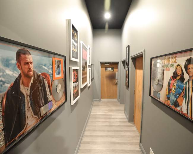 The image depicts a hallway lined with framed music awards and posters of artists on the walls, leading to a wooden door at the far end.