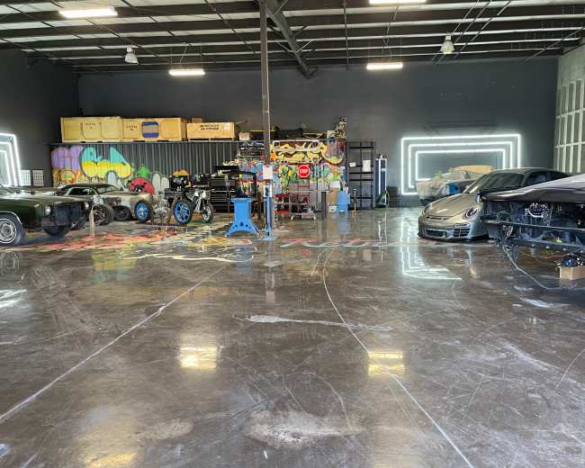 The image shows a spacious garage with several cars, including a classic car on the left and a modern car on the right, surrounded by graffiti art on the walls and polished concrete floors.
