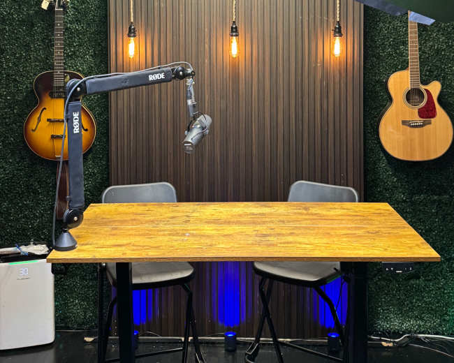 The image shows a recording studio setup featuring a wooden table, two chairs, hanging lights, and guitars mounted on a green wall backdrop.